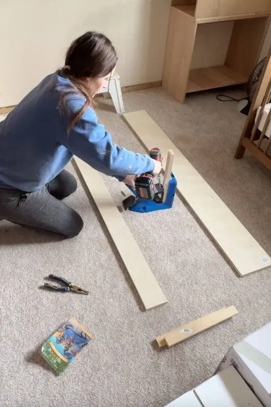 A DIYer kneeling on a carpeted floor using a pocket hole jig and a power drill to create angled holes in wooden boards for joinery. She is wearing a blue sweatshirt and gray leggings, working on a nursery project with various tools scattered around.